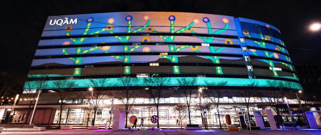 Photo du complexe des sciences de l'UQAM sur lequel une image colorée est projetée. On voit des arbres et une installation artistique à l'avant plan. Cette image illustre le campus de l'UQAM.
